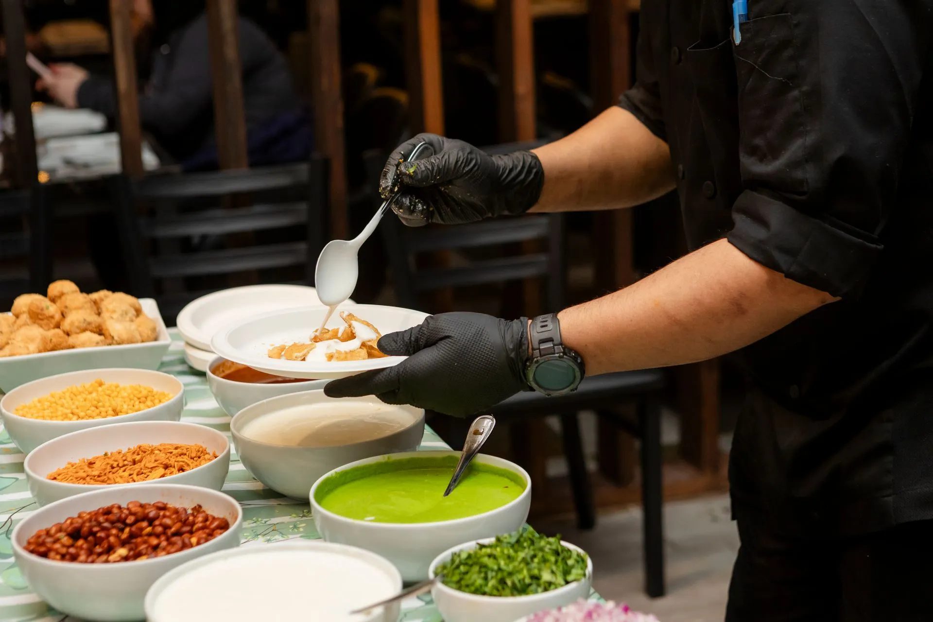 Chef plating Indian chaat at a restaurant counter lined with chutneys and toppings